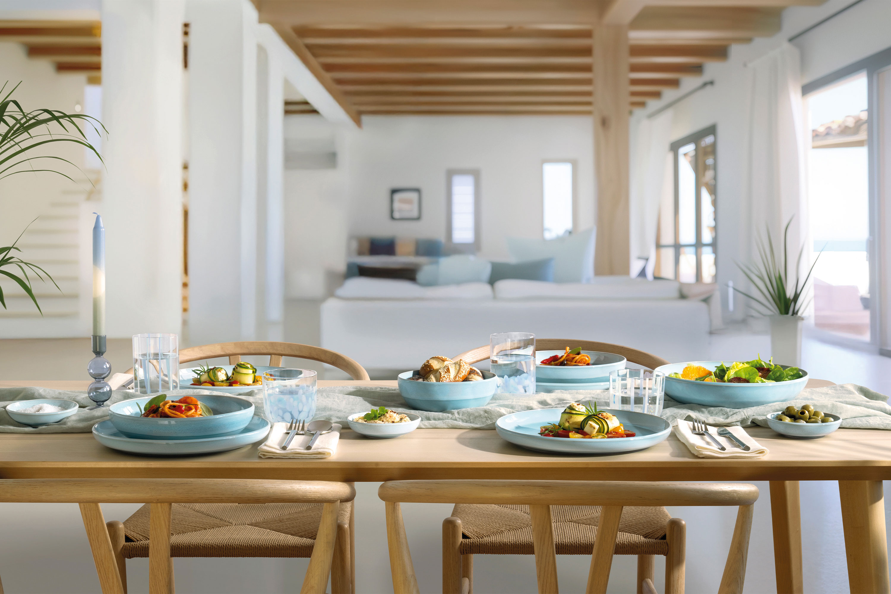 Un tavolo da pranzo in legno per quattro persone con piatti blu madreperla Aqua in una zona giorno luminosa e moderna.
