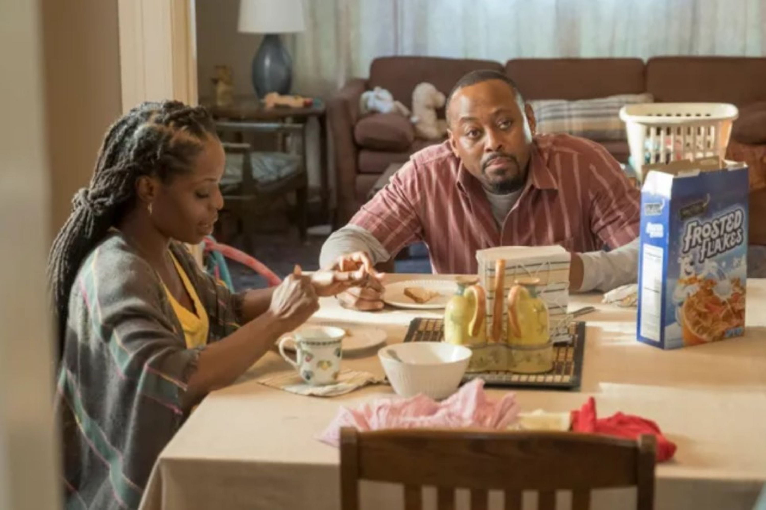 this-is-us-as-seen-on A couple sits at a dining table, with the man looking thoughtful and the woman eating breakfast. A cereal box and cups are on the table. The room feels cozy.