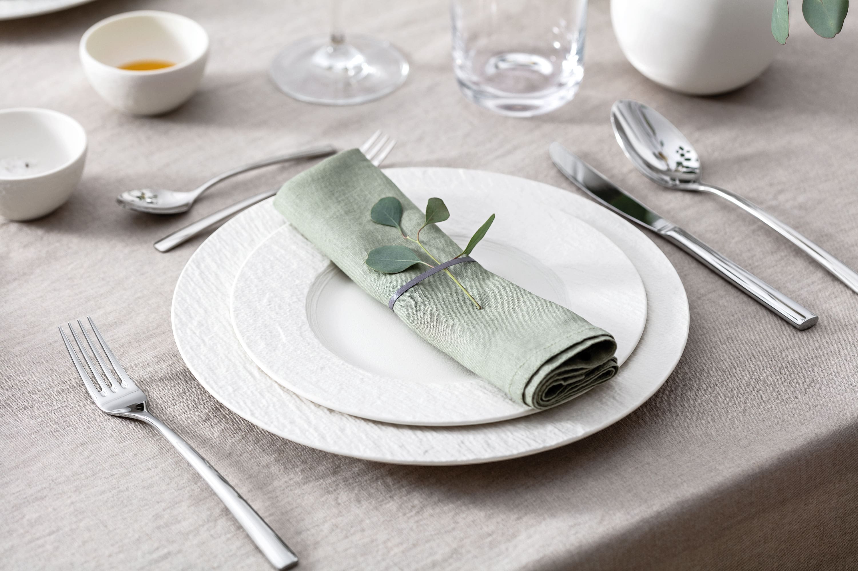 A set table with two white Manufacture plates, pastel green napkin, Piemont flatware and eucalyptus.