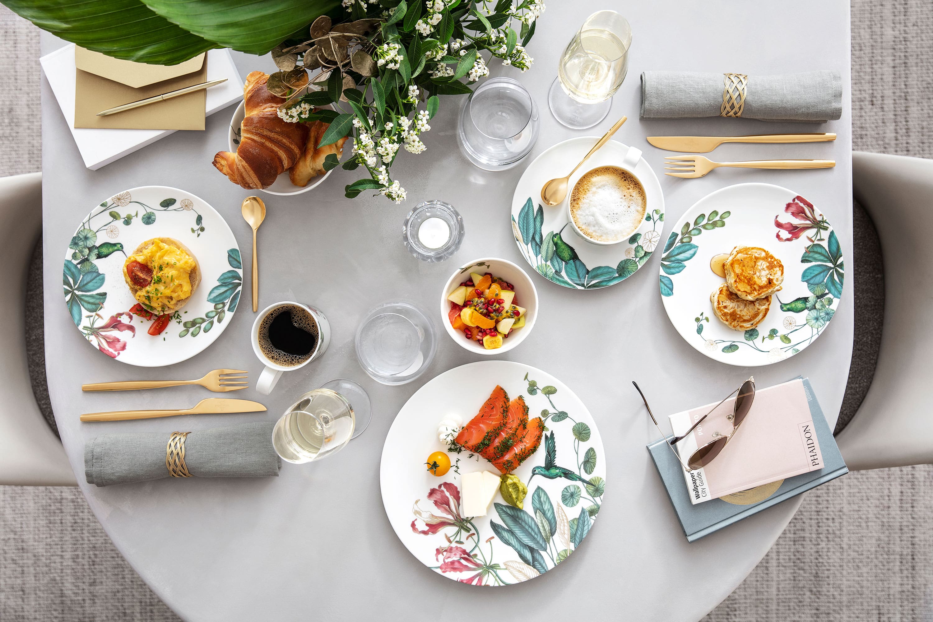 Une table de petit déjeuner Avarua Table dressée pour un repas avec des assiettes fleuries, des aliments, des boissons, des serviettes et un petit bouquet de fleurs pour décorer la table.