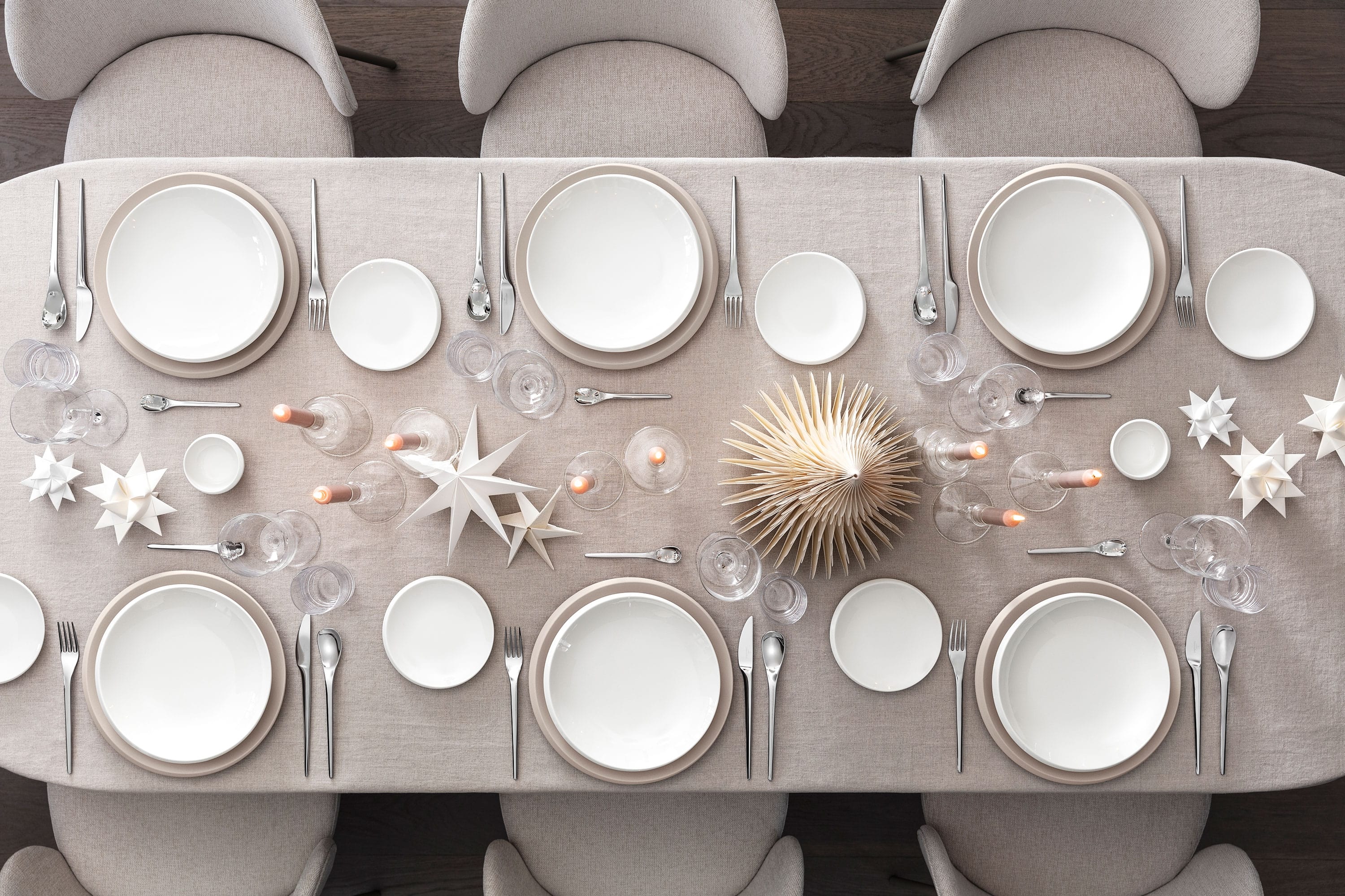Top-down view of a festive table setting for six featuring Villeroy & Boch’s NewMoon and NewMoon Beige collections: dinner plates, flat bowls, bread plates, and dip bowls. Accompanied by NewMoon wine glasses, tumblers, and 30-piece cutlery set. The table is styled with minimalistic holiday decor— white star ornaments and wooden Christmas trees—creating a warm, natural, and elegantly festive look.