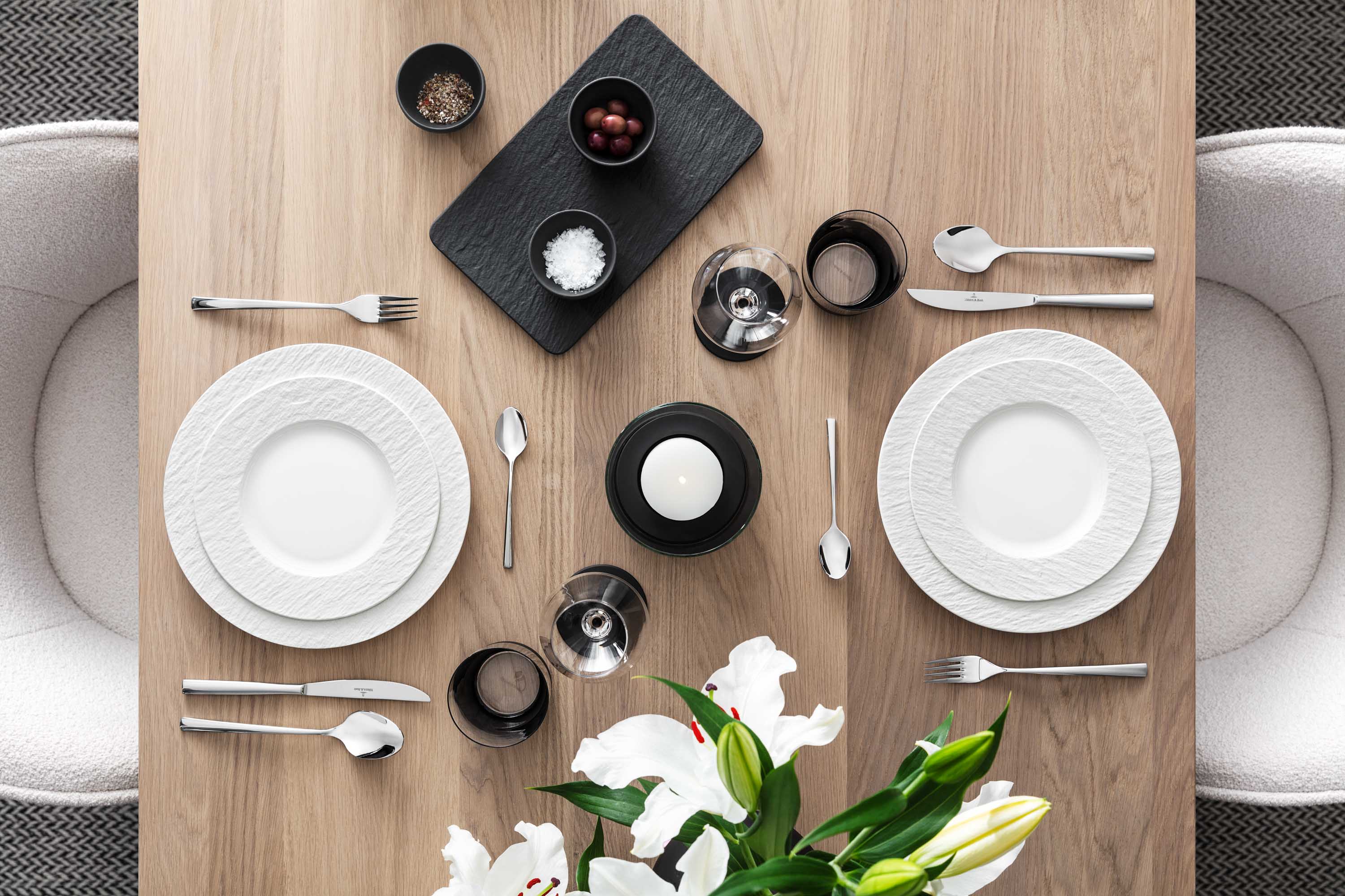 Elegant dining table with white Manufacture plates, Piemont flatware and decorative flowers on a wooden surface.