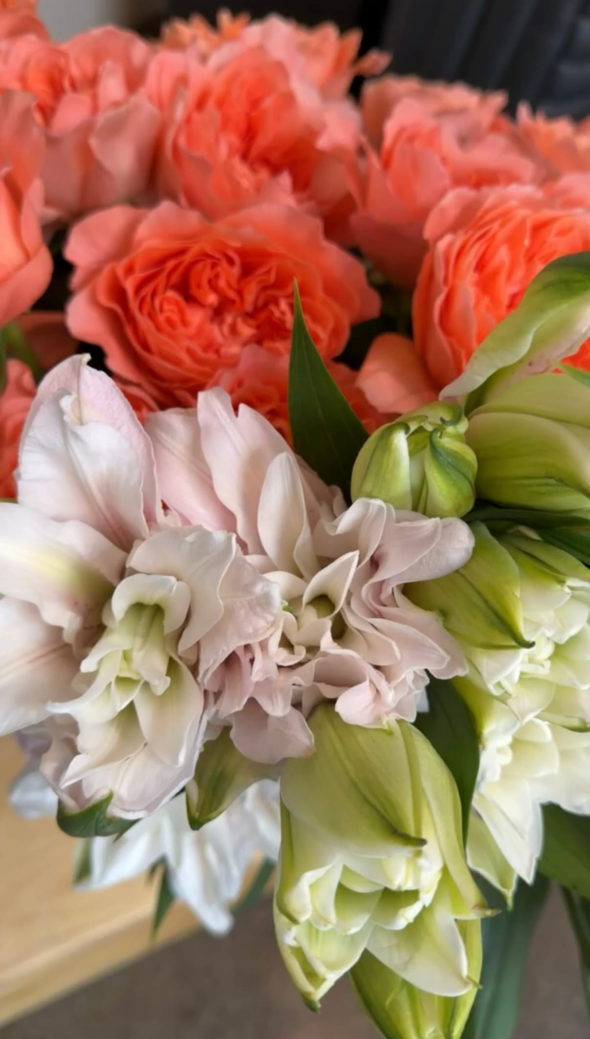 The Flowers Behind the Composition Close-up bouquet of coral roses, pale pink and white ruffled flowers, and green lilies, creating a lush, romantic, softly textured display.