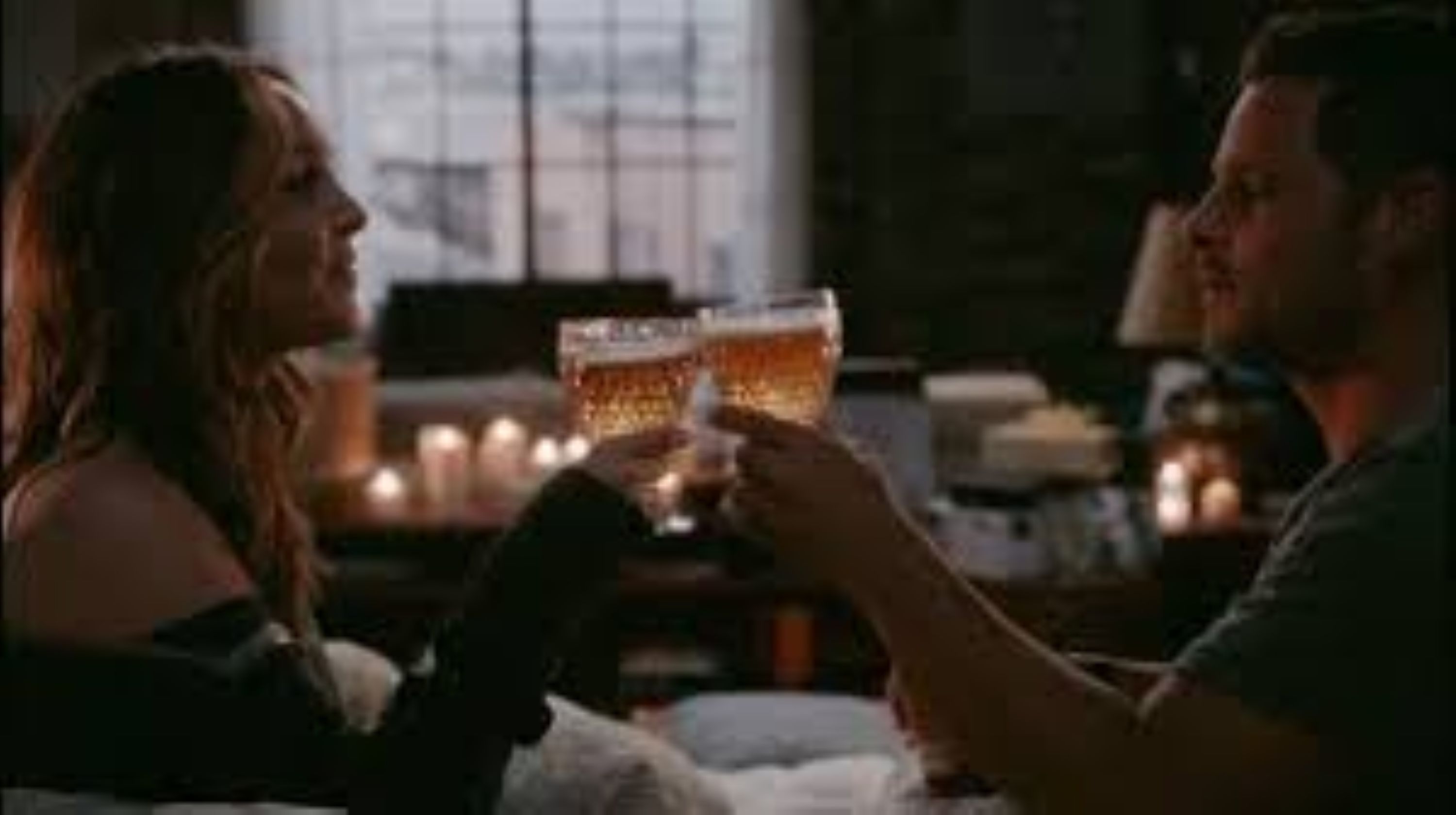 greys-anatomy-as-seen-on A couple toasts with beer glasses in a cozy, candlelit room, conveying warmth and intimacy. Dim lighting and a blurred window create a relaxed atmosphere.