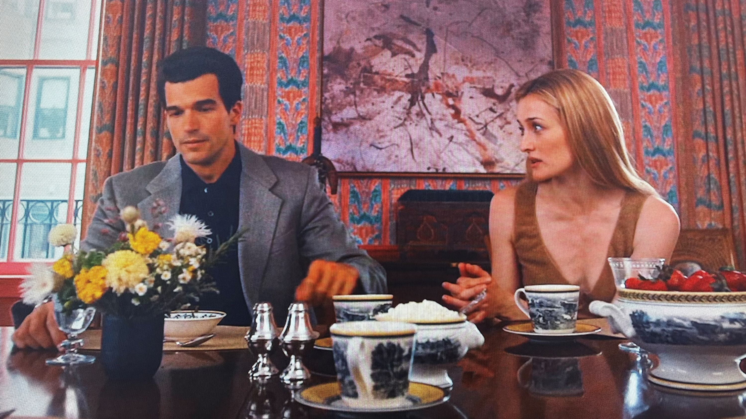 love-story-as-seen-on A man and woman seated at a dining table in an elegantly decorated room, engaging in conversation. The table is set with flowers, china, and silverware.