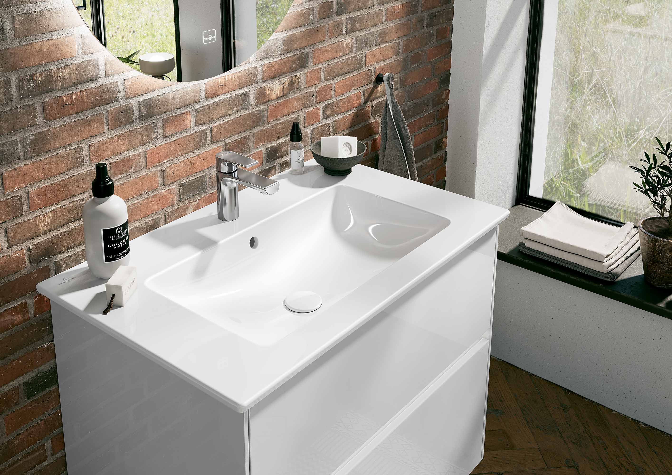 Un lavabo moderno con piano bianco, rubinetto cromato e specchio rotondo sopra. Sul top si trovano diversi articoli da bagno. Sullo sfondo, un muro di mattoni e una finestra.