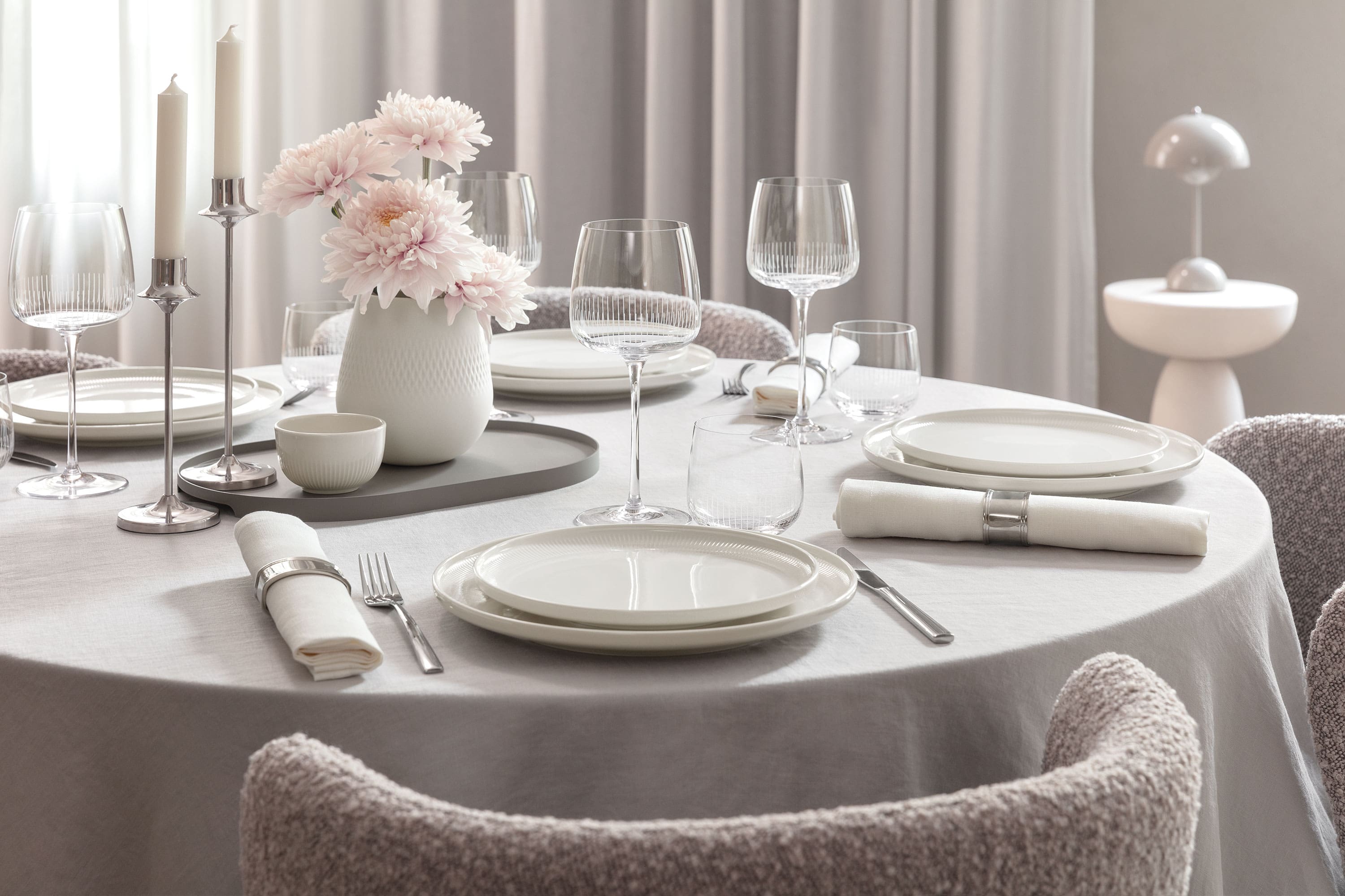 Tavolo da pranzo rotondo con piatti bianchi Afina, bicchieri Afina, posate, tovaglioli e un vaso con fiori rosa.