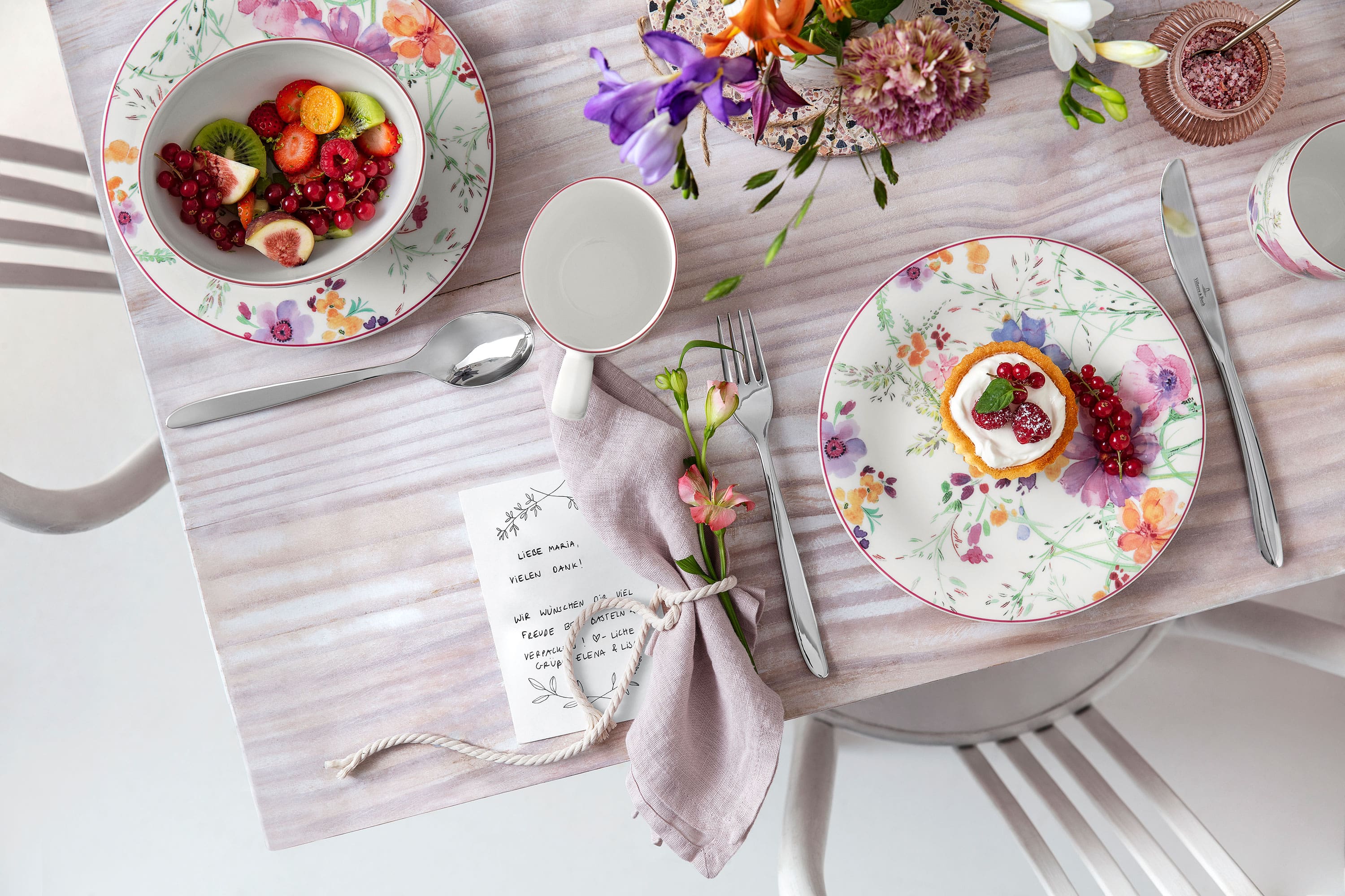 Ein mit Blumenmuster gedeckter Tisch mit Tellern, Besteck, einer Serviette, einem kleinen Dessert und einer Obstschale auf einer hellen Tischdecke.