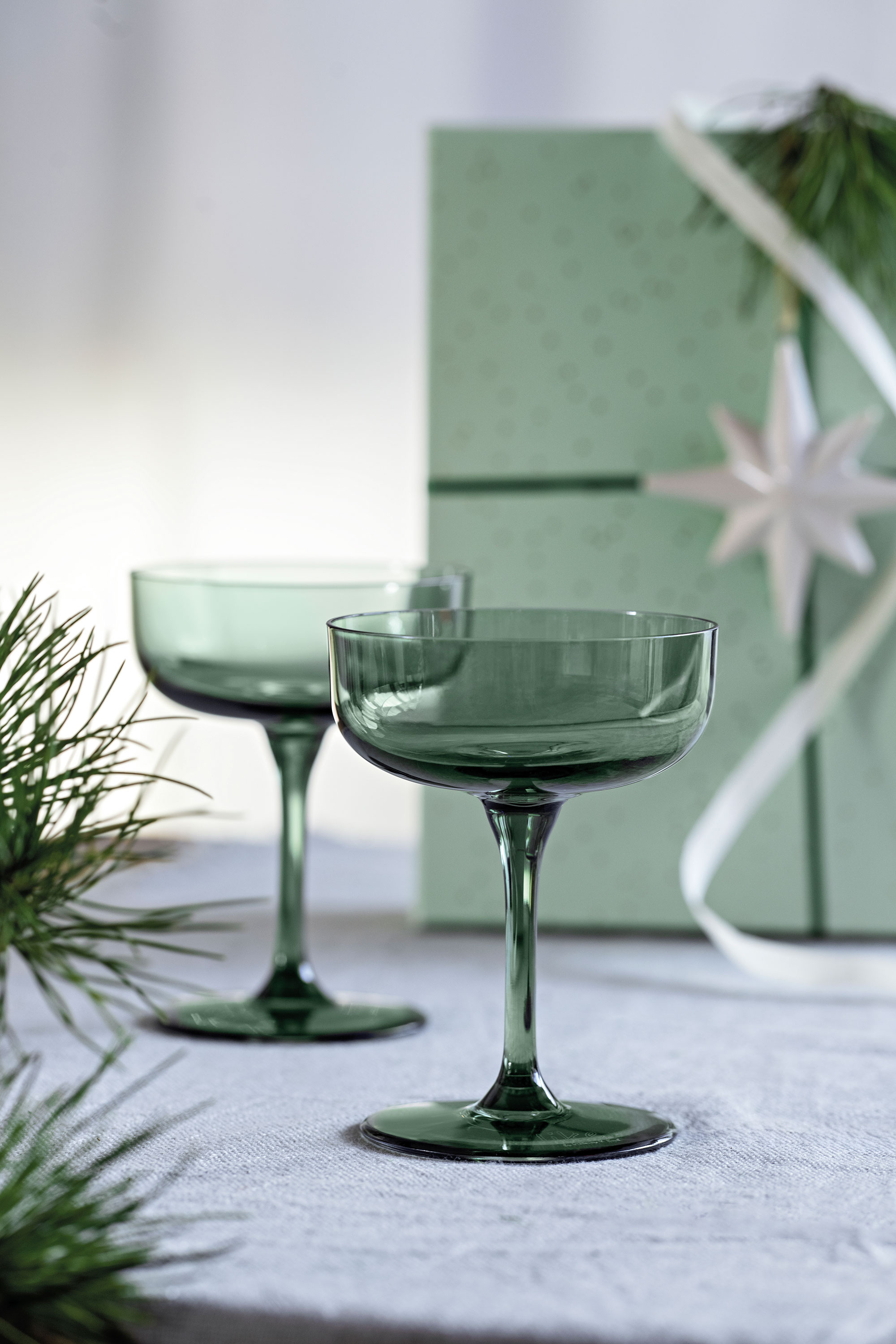 Two green cocktail glasses on a white surface with a green gift box and fir branches in the background. 