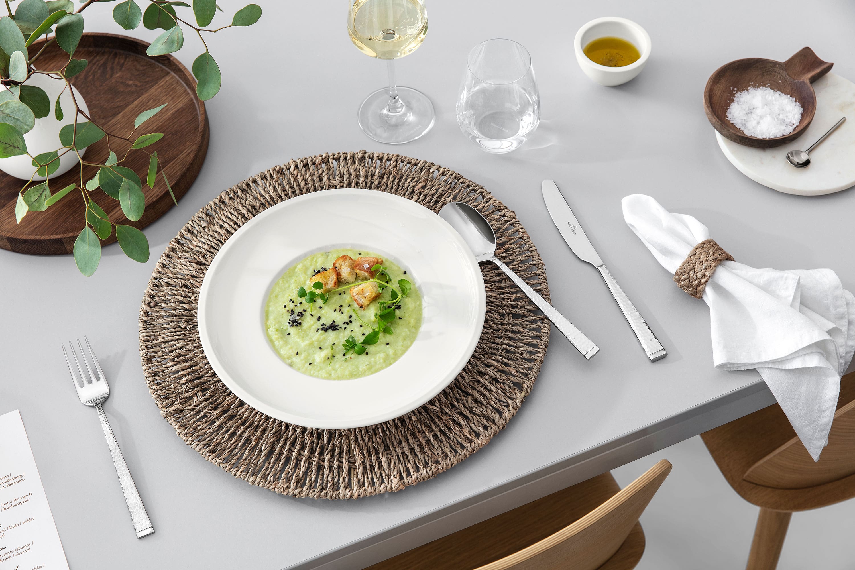 A dining table with Blacksmith flatware, an Artesano dish filled with soup, a glass of white wine and rustic table decorations.