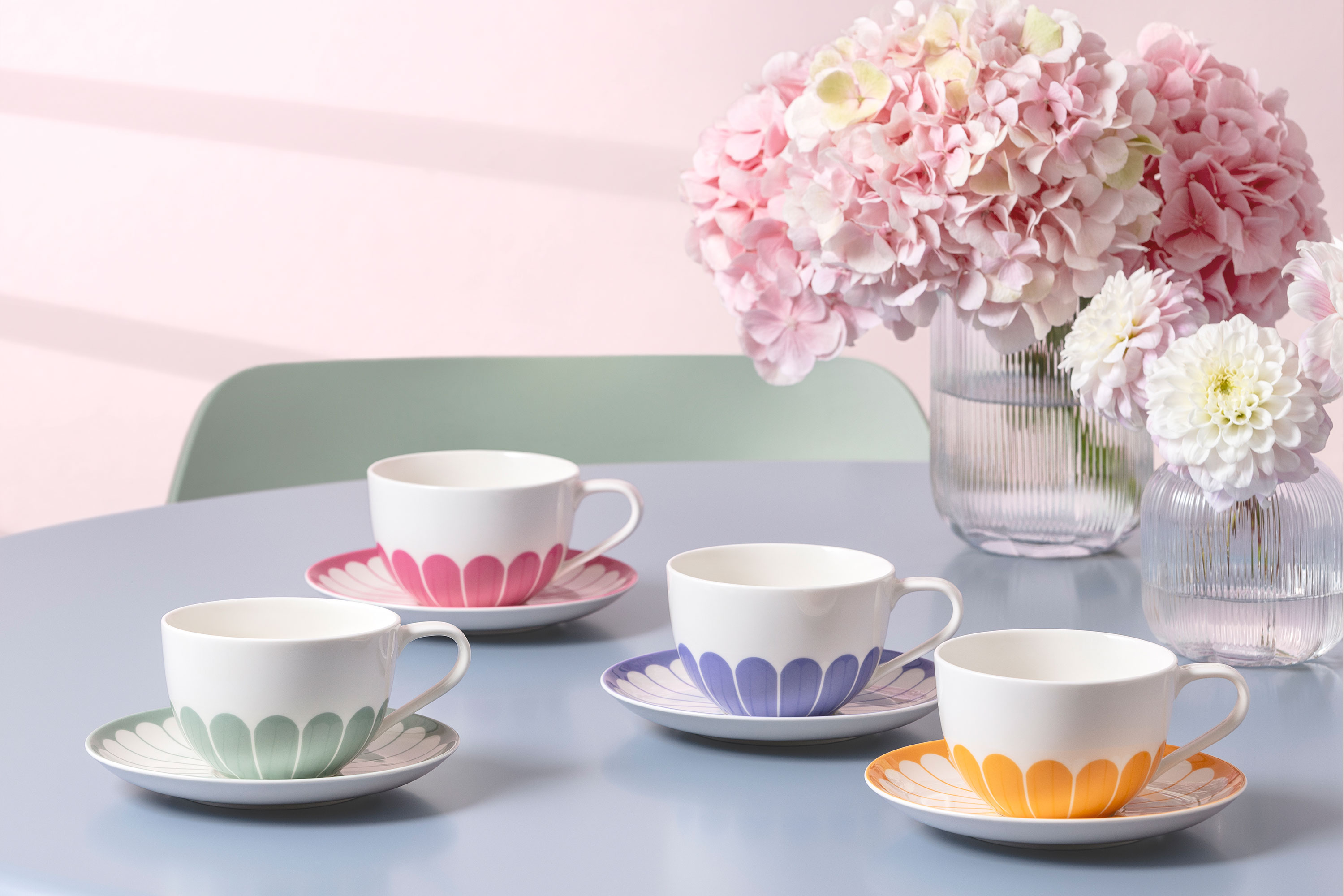 Quatre tasses à thé Fleur avec leurs soucoupes assorties, dans différentes couleurs pastel, sont disposées sur une table bleue à côté de vases contenant des fleurs roses et blanches.
